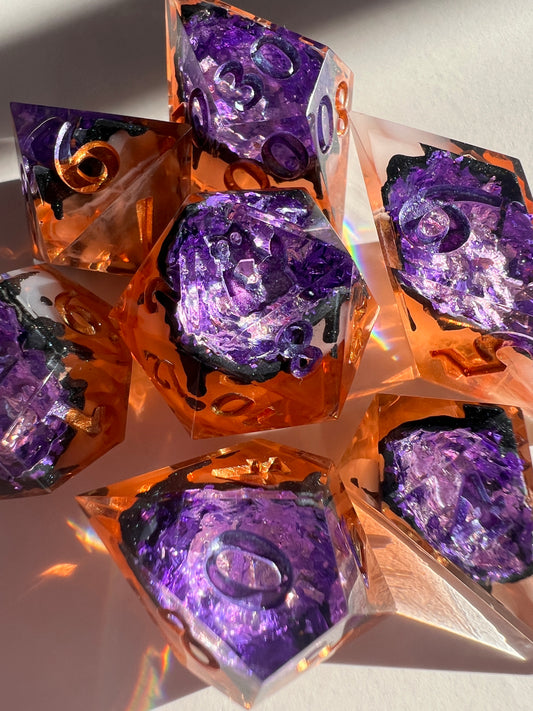 This spooky 7-piece geode-style dice set contains two different shades of purple foil to mimic crystals, a black inky rim, and an orange and cream-colored base.